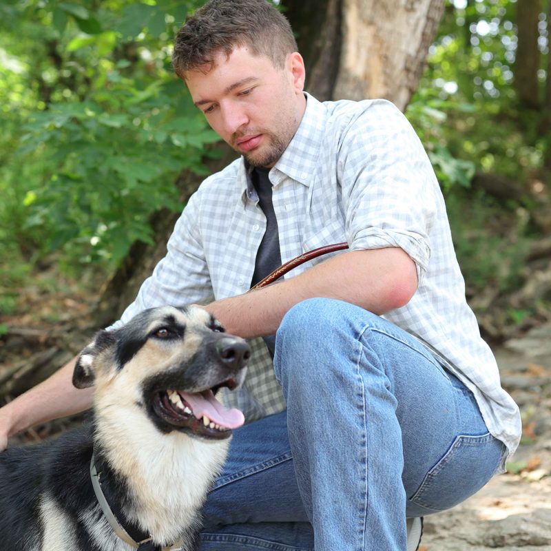 Rowan crouching next to a smiling German shepherd on a wooded trail